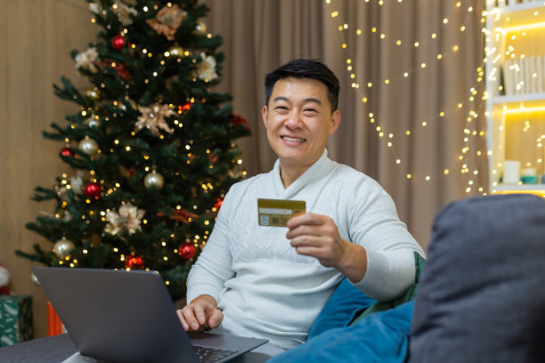 带着银行信用卡的男子微笑着，看着相机，带着笔记本电脑坐在家里客厅的沙发上，靠近圣诞树，快乐的亚洲购物者在网上商店为新年画像图片下载