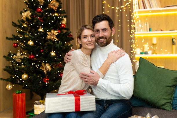 画像中，成熟的家庭大胡子男人和金发女人看着镜头拥抱在圣诞树旁，夫妇庆祝圣诞节和新年坐在客厅的沙发上，带着礼物图片下载