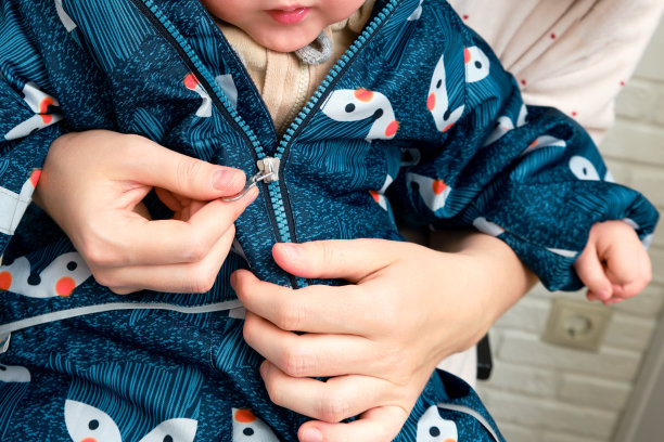 母亲给坐在家里走廊上蹒跚学步的婴儿穿上了一件蓝色的工作服。在寒冷的天气里，母亲给孩子穿保暖的衣服。一岁零三个月的孩子图片下载