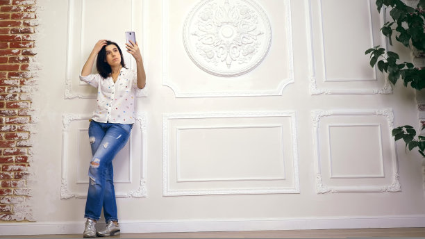 年轻女子，女孩，深色头发，在白色衬衫和牛仔裤，站在白色墙壁的背景装饰灰泥，与智能手机的肖像。看着屏幕，自拍，图片下载