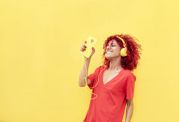 一头红色爆炸头的拉丁女子在黄色背景上跳舞，她拿着手机，戴着黄色耳机，享受着音乐图片下载