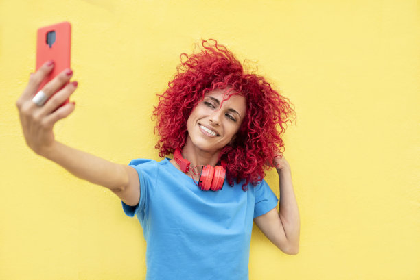 一名微笑的拉丁妇女，一头红色的爆炸头，穿着蓝色的t恤，脖子上戴着红色的耳机，背景是黄色，正在自拍图片下载