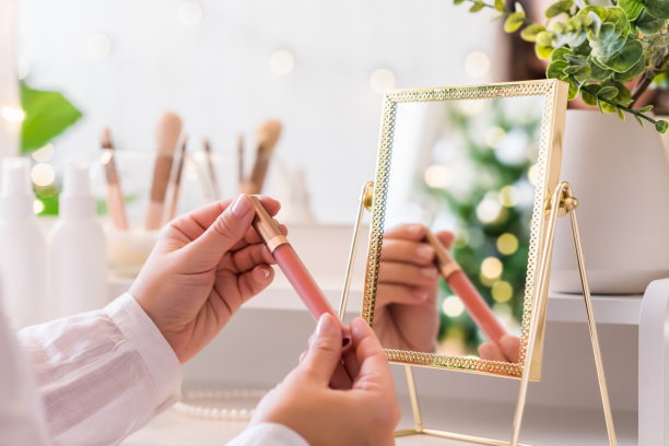 一位女士正在涂口红，坐在化妆台旁边，旁边放着化妆配件。圣诞树的倒影，桌上镜子里的灯泡。图片下载