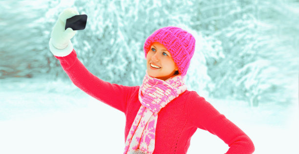 冬天的肖像，快乐的微笑年轻女子用智能手机自拍，穿着针织毛衣，帽子和围巾在公园的雪景树背景图片下载