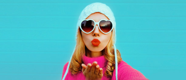 冬天的画像，美丽的年轻女子伸出手用智能手机自拍，穿着针织毛衣，白色帽子，蓝色背景的心形太阳镜图片下载