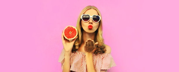 夏天的肖像美丽的年轻女子与一片多汁的葡萄柚吹她的嘴唇送甜蜜的飞吻戴心形太阳镜在粉红色的背景图片下载