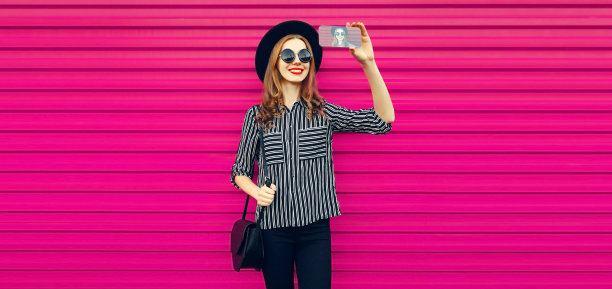 肖像快乐微笑的年轻女子与智能手机自拍戴黑色圆帽在粉红色的背景图片下载