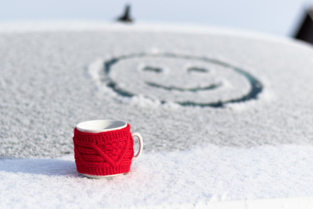 在一个霜冻明媚的冬日，一个红色的马克杯和一杯热饮放在积雪的汽车行李箱上。在汽车的后窗上，画着一张笑脸。有选择性的重点。图片下载