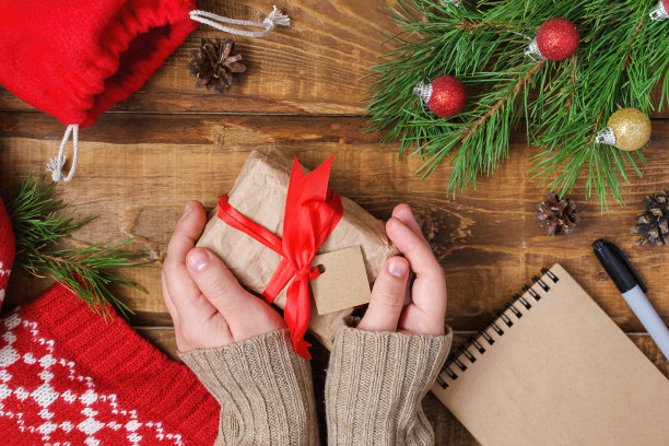 俯视图平躺在一张木桌上，女性在一件针织毛衣手里拿着一个礼物与红色缎带蝴蝶结的形式的框架躺着绿色杉树树枝红色图案格纹和一个记事本。图片下载