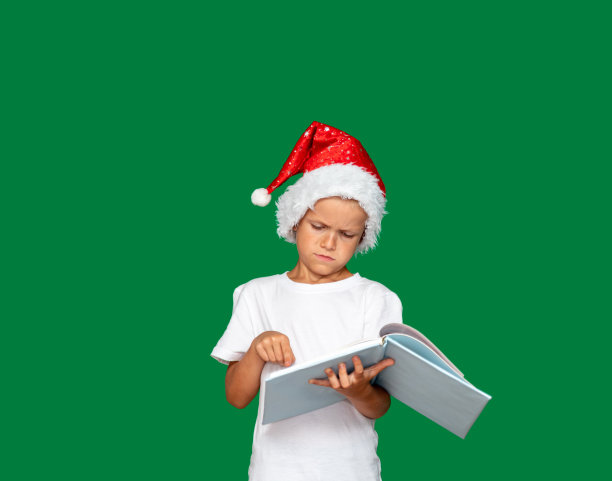 戴着圣诞老人帽子的男孩愤怒地盯着一本打开的书。绿色背景，留有文字空间。有选择性的重点。为关于儿童、节日、圣诞节的文章和广告拍照。图片下载