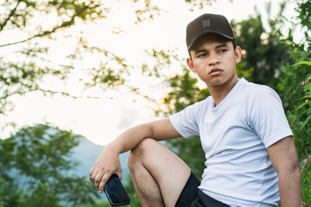 年轻的拉丁人，坐在山顶上看着美丽的自然，背景是金色的日落。男孩手里拿着手机。黑色帽子和白色t恤。图片下载