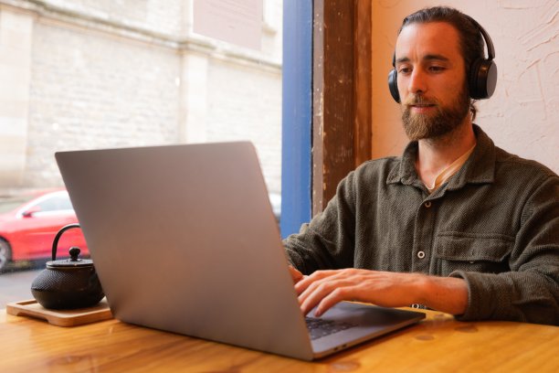 胡子男坐在靠窗的地方，戴着耳机，用电脑工作图片下载