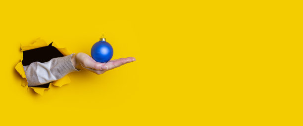 女人的手拿着一个黄色背景的蓝色装饰圣诞球。横幅图片下载