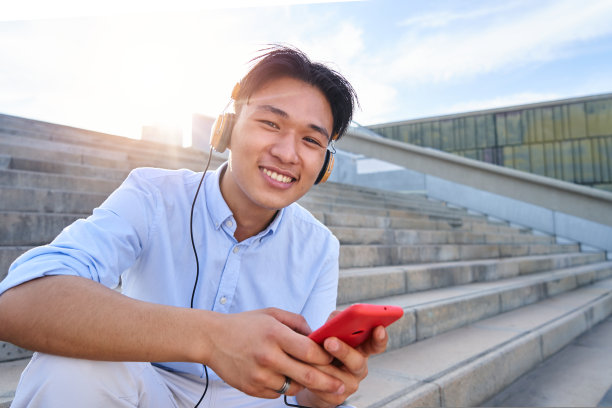 一个男孩用耳机听音乐，坐在楼梯上看着相机的快乐肖像。图片下载