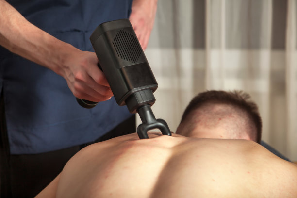 在健身房的医务室用枪敲击按摩背部肌肉健壮的男子。按摩师做按摩练习。运动身体肿块再生的打击疗法。损伤康复的概念图片下载