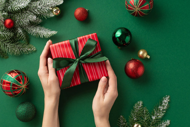 平安夜的概念。第一人称俯视图照片的妇女的手拿着红色礼盒丝带蝴蝶结在绿色金红色小饰物球和松树树枝在霜冻孤立的绿色背景图片下载