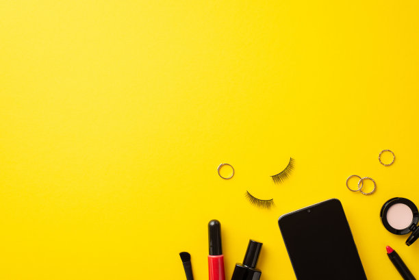 网络星期一的概念。俯视图照片的智能手机化妆品指甲油假睫毛眼影唇眼线，光泽刷和金戒指在孤立的黄色背景与空白图片下载