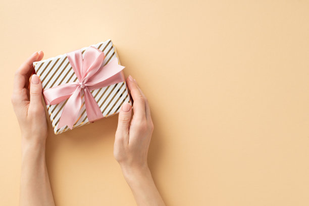 圣诞庆祝概念。第一人称俯视图照片的女孩的手拿着时髦的礼品盒与粉色丝带蝴蝶结孤立的彩色米色背景与copyspace图片下载