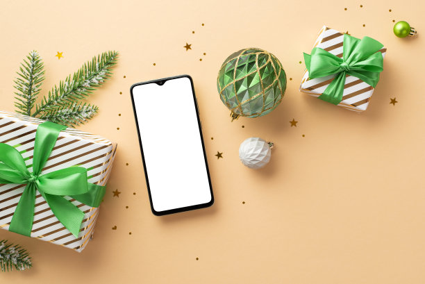 新年的概念。俯视照片的智能手机白色和绿色的装饰物礼盒松枝在白霜和纸屑孤立的米色背景与copyspace图片下载
