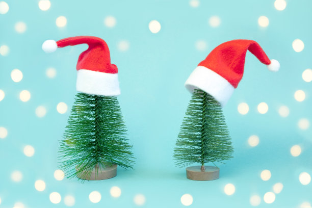 两棵戴着红色圣诞帽的小圣诞树。装饰人造杉树。漂亮的迷你圣诞树，蓝色背景上有圣诞老人帽。圣诞假期的概念。极简主义。图片下载