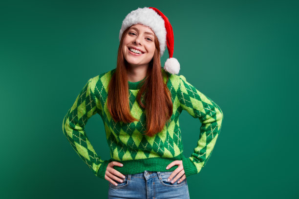 美丽的年轻女子戴着圣诞帽，手拉在臀部，微笑在绿色的背景图片下载