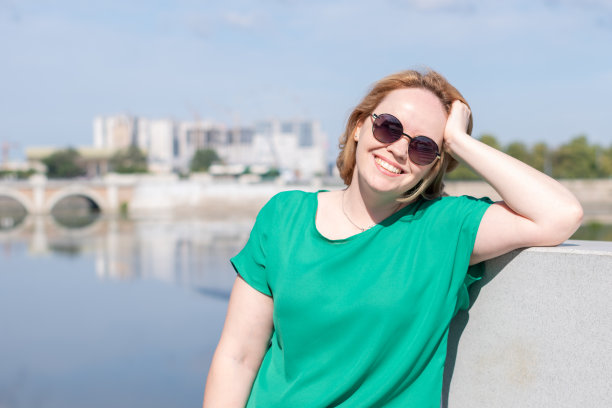 一个戴着墨镜，穿着绿色上衣的女孩站在河边用手抓着头发的肖像，复制空间。图片下载