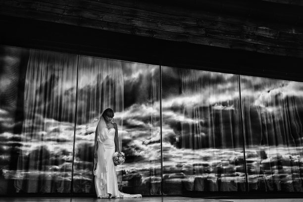 黑色和白色的照片，年轻的新娘在白色长裙和手捧花束在户外的大镜子窗与多云的天空背景。结婚的那一天图片下载