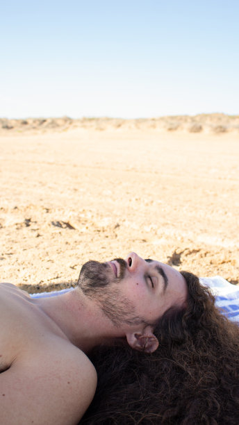 垂直拍摄的年轻迷人的男子长卷头发躺在沙滩毛巾图片下载