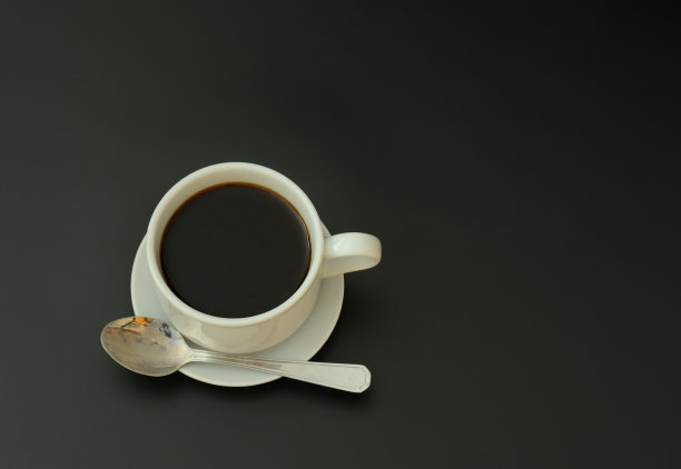 一杯热黑咖啡放在碟子上，勺子放在黑色背景上。图片下载