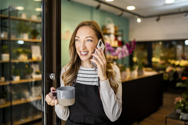 微笑的年轻花匠在花店用智能手机拿着一杯咖啡图片下载