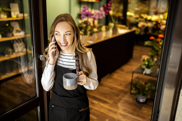 年轻的花匠在花店用智能手机端着一杯咖啡图片下载