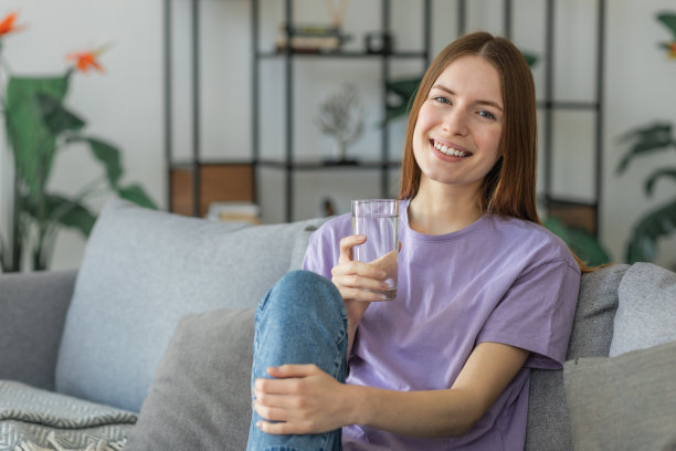 健康的生活方式理念。美丽的年轻白人妇女拿着一杯新鲜的水坐在家里的沙发上，微笑着友好图片下载