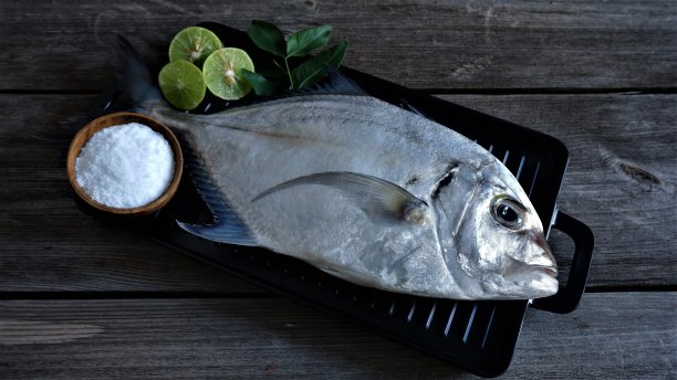 新鲜的鲳鱼装在黑色托盘里，配上咖喱叶、一片酸橙和盐，放在木桌上。Alectis Ciliaris。图片下载