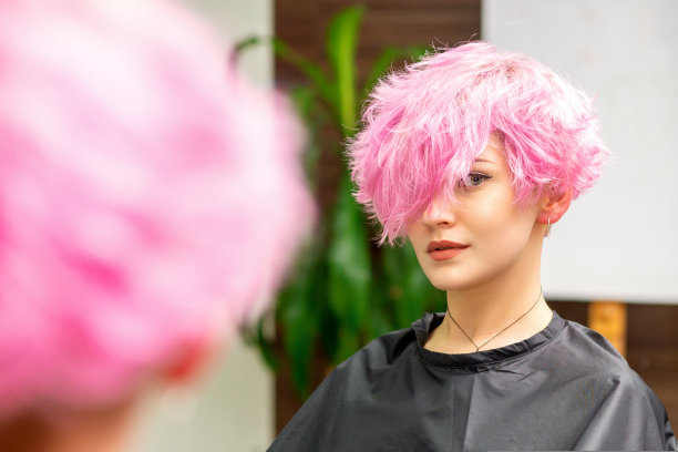在一家美发店里，镜子里的倒影里，梳着粉色短发的美丽白人年轻女子正对着镜头。图片下载