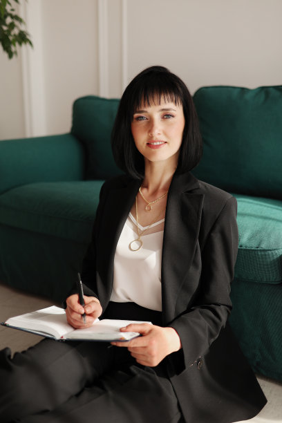 微笑的女商人在笔记本上写字，看着镜头。深色头发的女人坐在绿色沙发旁边，穿着黑色西装和高跟鞋。室内种植绿色植物。居家办公图片下载