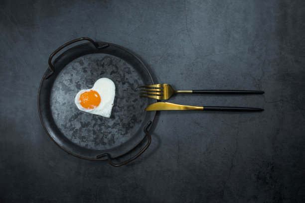 横幅。自制煎蛋在一个老式的平底锅和时尚的餐具在黑暗的背景。心形的蛋黄。从上面看。文本的复制空间。图片下载