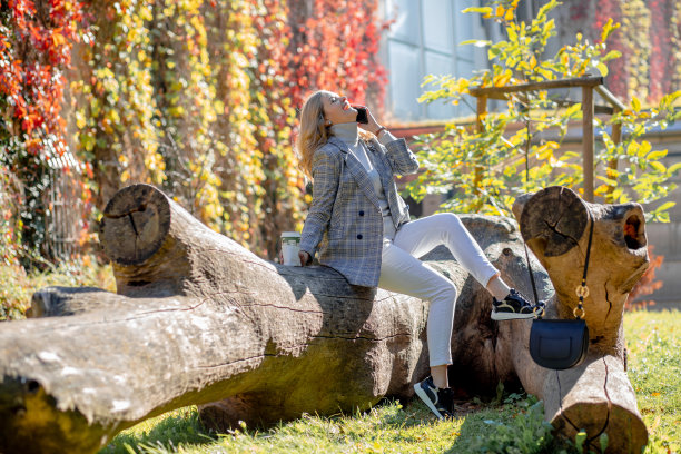 秋日阳光明媚，一位美女坐在秋园的大倒树上，仰着头漫不经心地笑着。积极的情感。享受的时刻。咖啡休息。图片下载