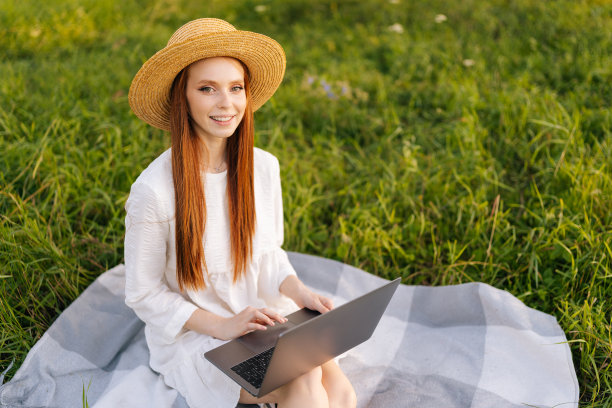肖像快乐优雅的年轻红发女子戴草帽和白色连衣裙坐在摆姿势在格纹与笔记本电脑看着相机。图片下载