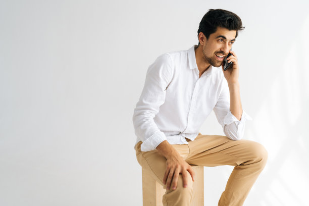 工作室肖像的英俊微笑的男人在休闲衬衫上说话的智能手机，微笑和享受移动通话坐在白色孤立的背景。图片下载