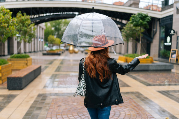 背影依稀可辨的优雅女子，戴着时尚的帽子，打着透明的雨伞走在美丽的城市街道上，用手感受着雨。图片下载