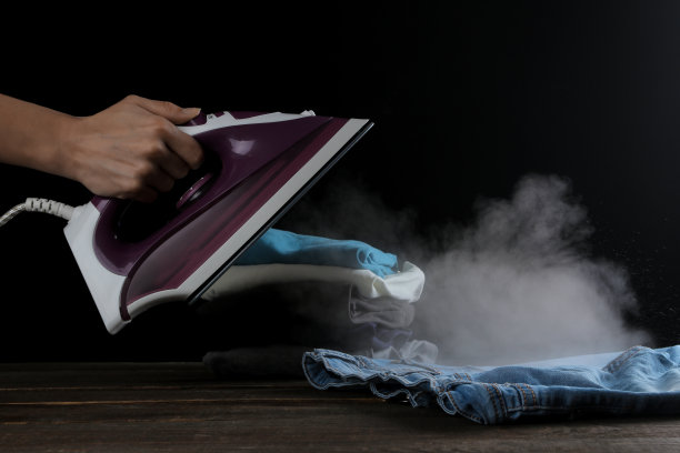 这个女孩在黑色背景的衣服上散发淡紫色熨斗的蒸汽。熨烫衣服。家用电器。图片下载