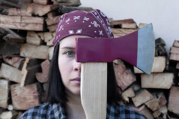 一个严肃的年轻成年妇女站在镜头前，拿着一把斧头在她的面前。在背景上收割柴火，准备迎接寒冷的冬天，以度过能源危机。图片下载