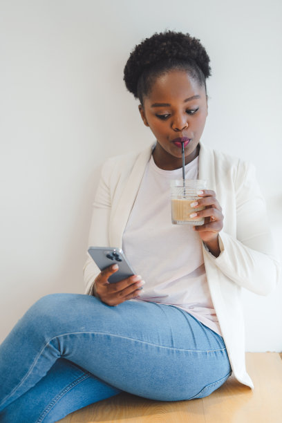 非洲裔美国妇女的肖像与非洲发型一边喝冷饮一边看手机。迷人的美丽女孩。时尚模特。美丽的肖像。图片下载