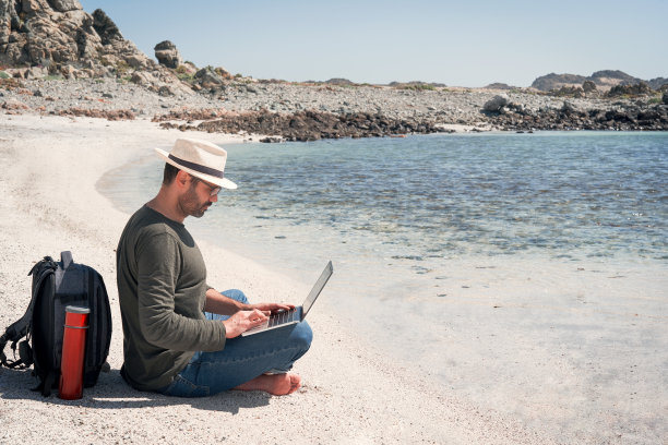 成熟的拉丁男性坐在海滩的岸边，悠闲地摆弄着笔记本电脑图片下载
