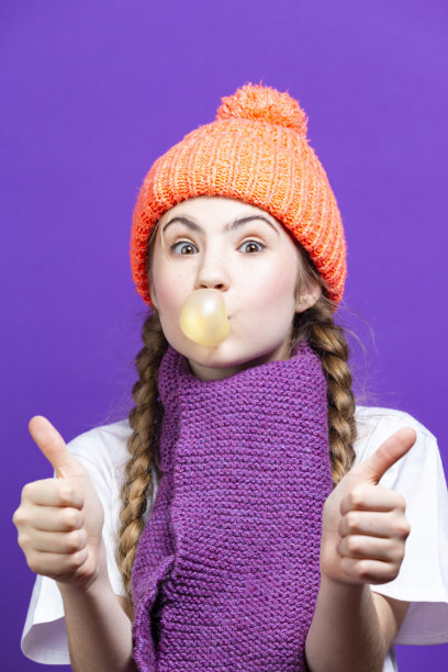 现代青年观念。一个戴着珊瑚色针织季节帽的女孩，在紫色背景下，把口香糖泡放在面前，竖起大拇指图片下载