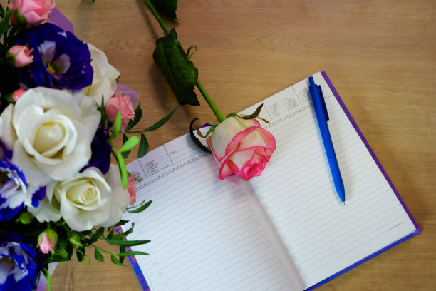 Flatlay。记事本、日记本和一支笔放在木桌上，旁边是一束五颜六色的玫瑰。工作空间与日记和粉白色的花在木制背景。俯视图图片下载