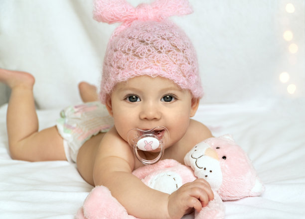 小婴儿戴着粉红色的帽子，穿着尿布，躺在床上，手里拿着玩具，吮吸着奶嘴图片下载