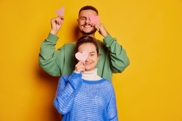 美丽的年轻夫妇的肖像，男人和女人摆姿势与纸心隔离在黄色背景。快乐的庆祝活动图片下载