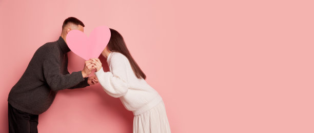 可爱的年轻夫妇的肖像，男人和女人接吻背后的纸心，摆出孤立的粉色背景。带有复制空间的传单图片下载