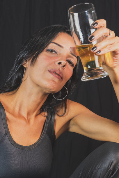 肖像年轻女子在黑色t恤喝啤酒从玻璃杯。在黑色的演播室背景下。图片下载